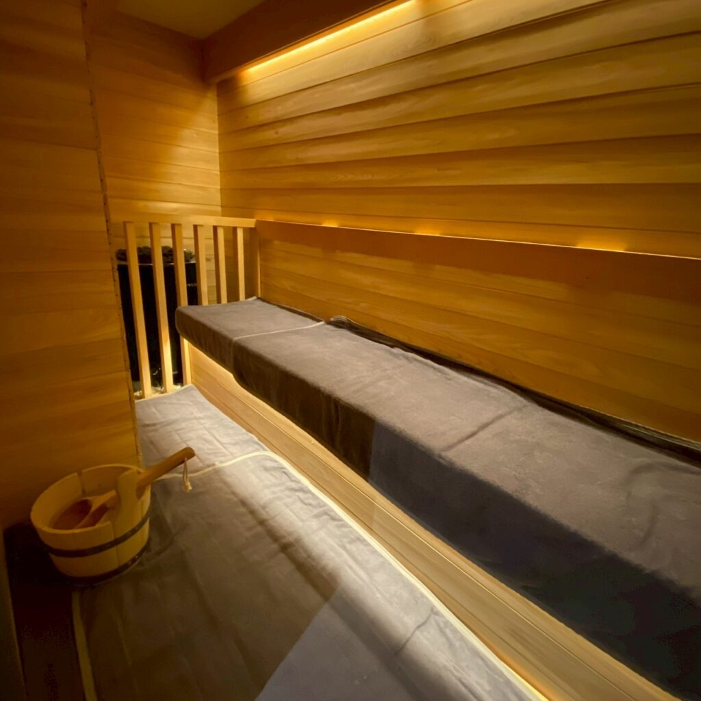 木材で仕上げられたサウナルーム。間接照明が温かく灯り、黒いサウナマットが敷かれた落ち着いた空間。