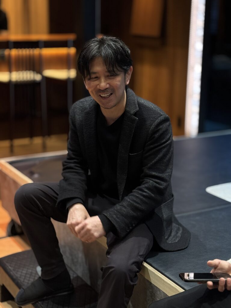 ジム内のスペースに座り、リラックスした様子で笑顔を見せる男性。背後には木目のインテリアが広がっている。