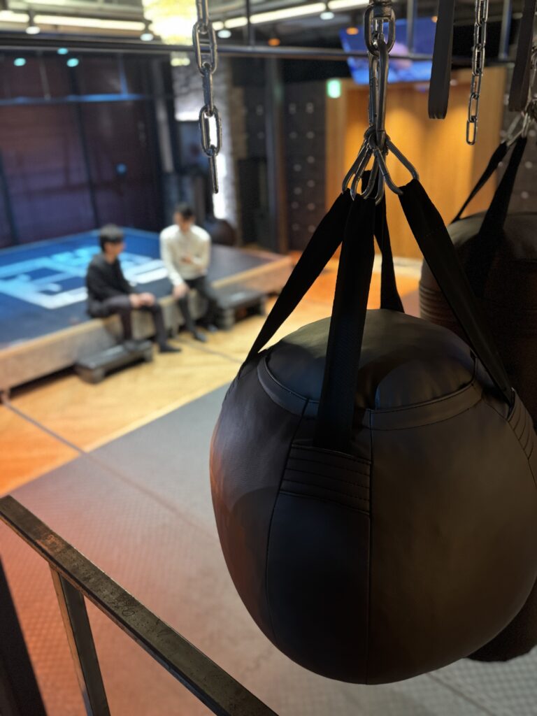 吊り下げられた黒いサンドバッグの奥で、リング横に座った2人の男性が会話している様子。ジム内の照明が温かく空間を照らしている。
