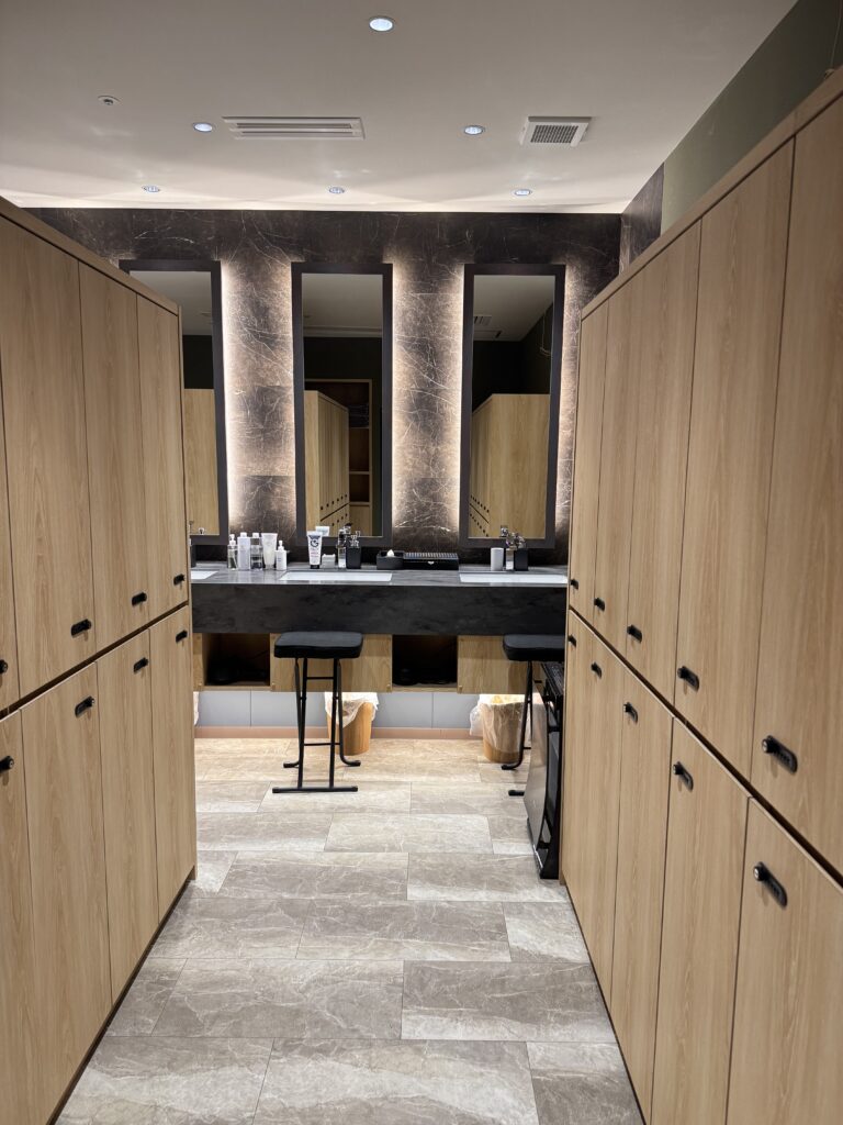 木目のロッカーが並ぶOYG銀座ジムのパウダールーム。奥にはライトアップされたミラーとカウンターがあり、ケア用品が整然と並んでいる。