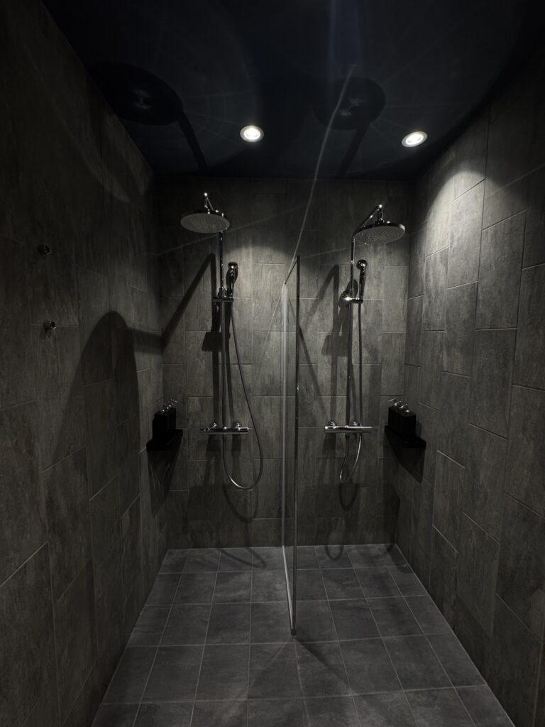 暗めの石材タイルで統一されたシャワールーム。2つのレインシャワーが向かい合うように設置され、落ち着いた照明が空間を照らしている。