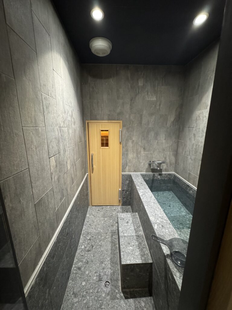 グレーの石材で仕上げられた静かな浴室空間。奥には木製扉のサウナルームがあり、手前には水を満たした浴槽が設置されている。