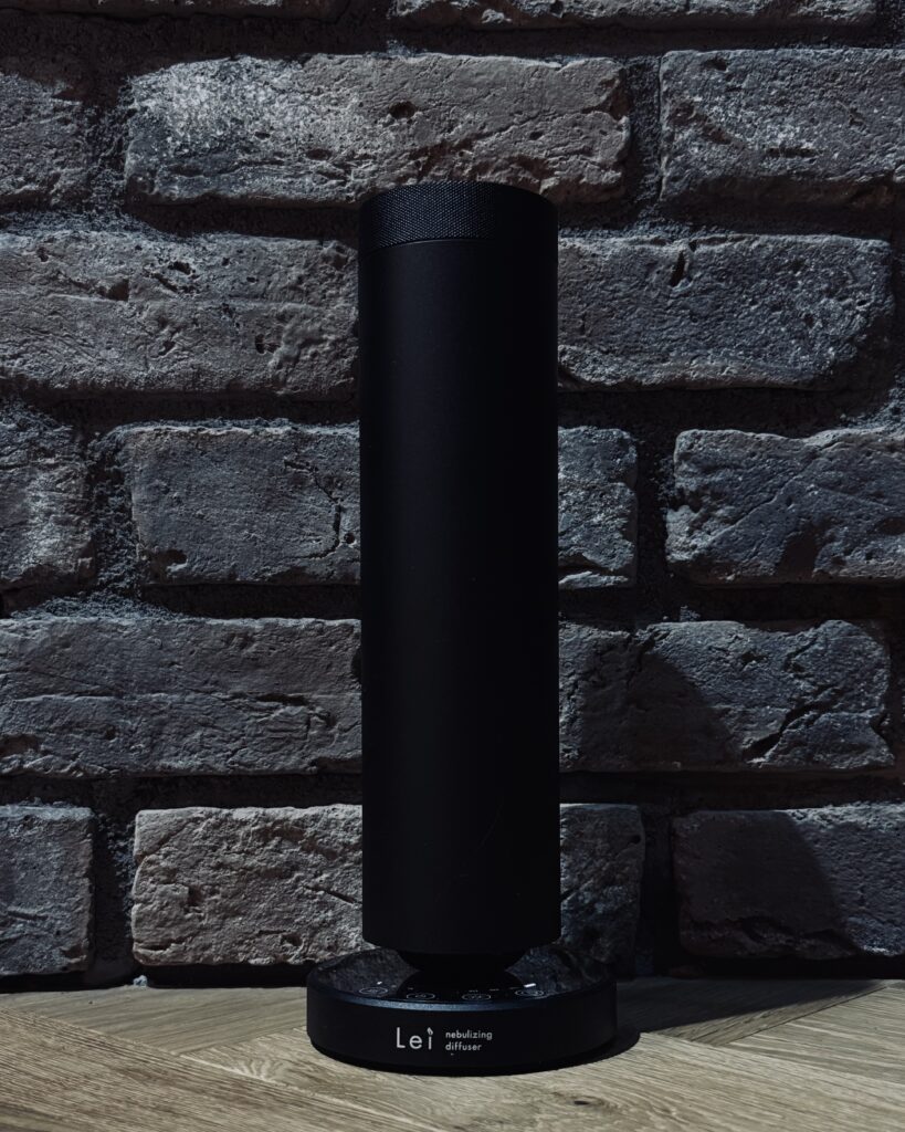 レンガ壁の前に静かに佇む黒いLei業務用アロマディフューザー。陰影の中で存在を主張せず、空間に香りを満たしている。