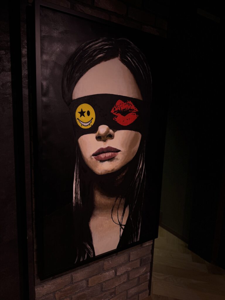目元を黒いアイマスクで覆われ、スマイルマークと唇のイラストが描かれた女性の肖像アート。暗い壁面にディスプレイされている。
