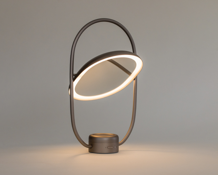 Featuring an elliptical ring frame and an angled circular LED light, this modern portable product combines lighting, mirror and aroma diffuser functions. It emits a warm glow against a neutral backdrop.