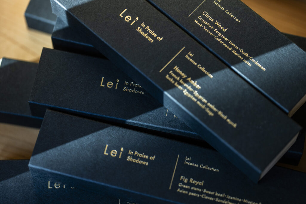 Stacked matte-black Lei “In Praise of Shadows” incense boxes with gold-foil lettering, showing scents including Citrus Wood, Honey Amber, and Fig Royal.