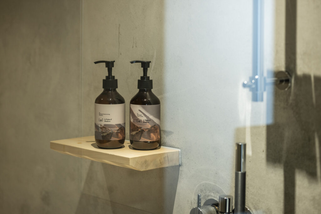 Two amber pump bottles of Lei KUON Perfumed Moisturizing Shampoo and Perfumed Liquid Soap on a small wooden bathroom shelf, lit by soft daylight.