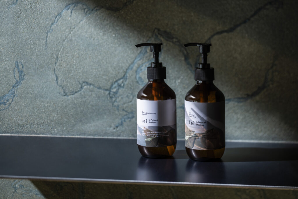 Two amber pump bottles of Lei KUON Perfumed Moisturizing Shampoo and KUON Perfumed Liquid Soap on a dark shelf against a textured stone wall.