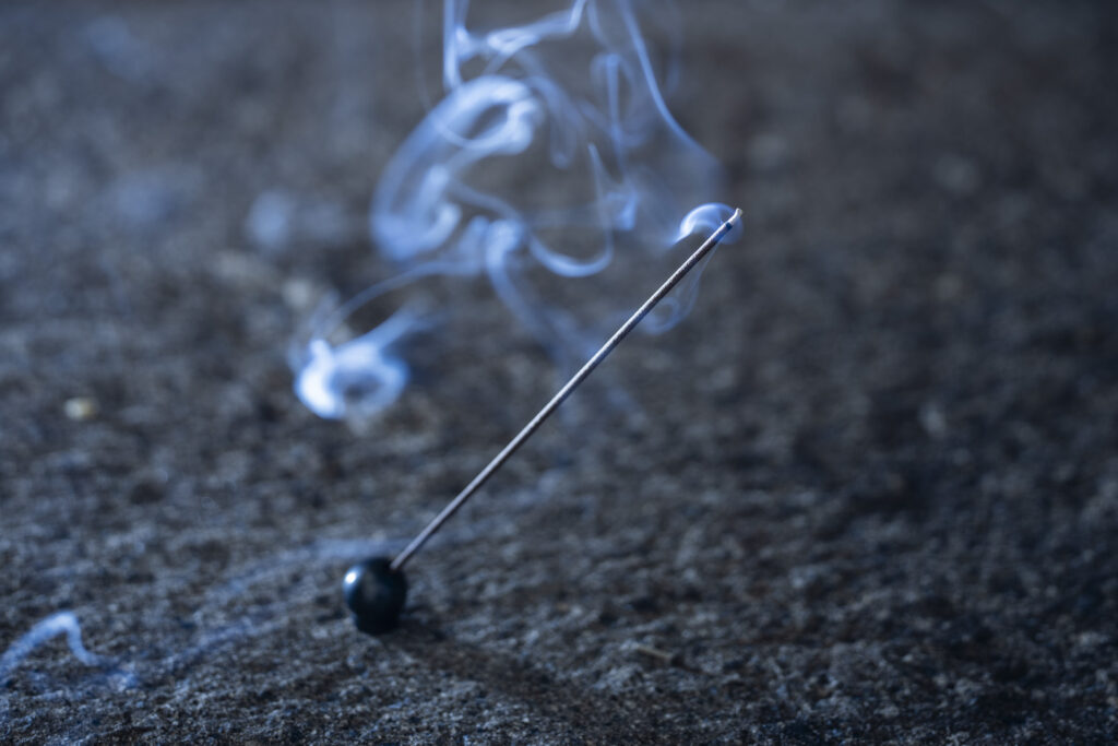 A lit incense stick resting on a small black holder, with pale smoke curling softly over a dark textured surface.
