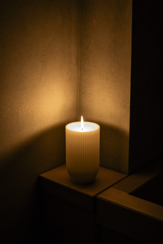A Japanese Lantern Candle in a ribbed holder placed on a small wooden shelf, casting warm light and deep shadows against a textured wall.