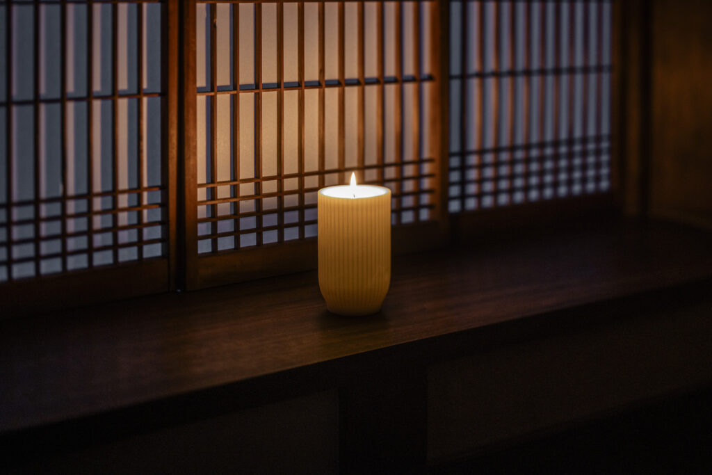 A lit candle in a ribbed holder placed on a wooden ledge in front of a shoji-style lattice screen.