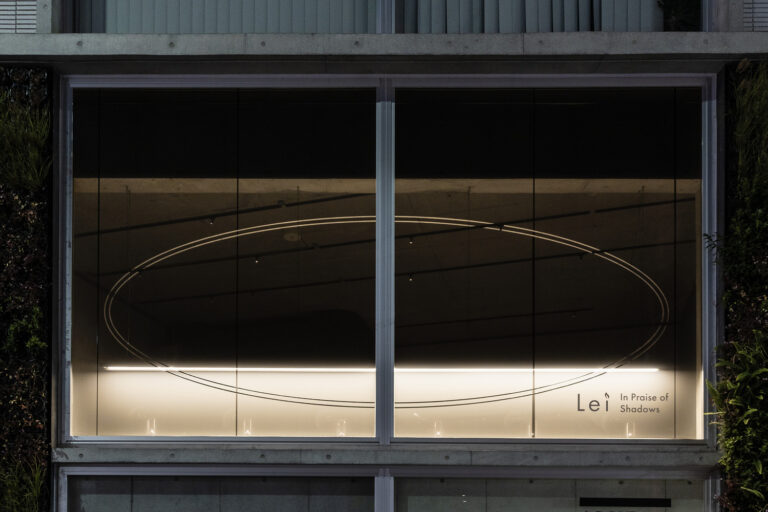 The exterior of Lei In Praise of Shadows Flagship Store at night, with an oval outline and the store’s logo softly illuminated on the large glass window.