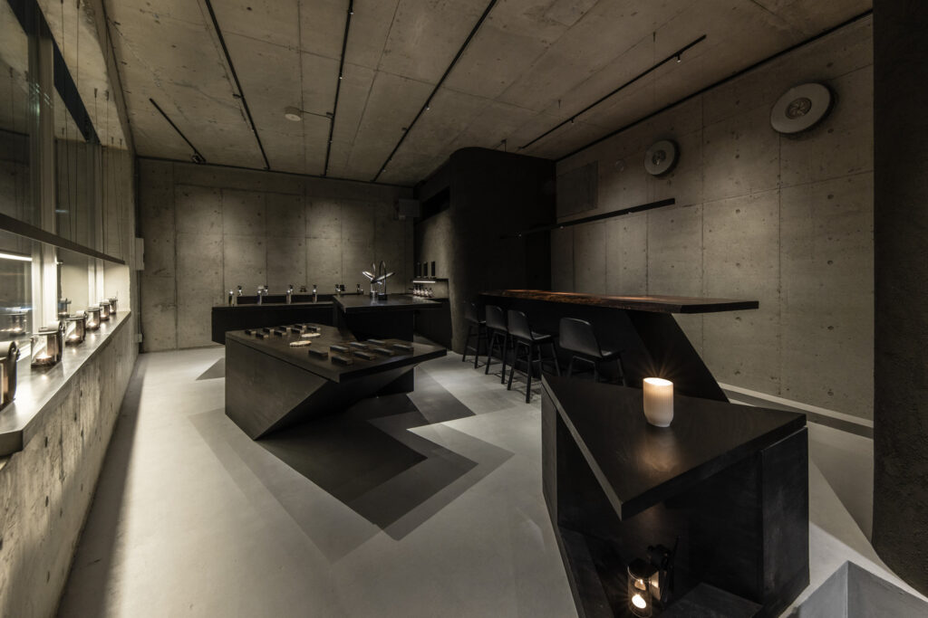 Candlelit interior of Lei In Praise of Shadows Flagship Store, featuring concrete walls, minimal black furniture, and a bar counter designed for light and shadow.