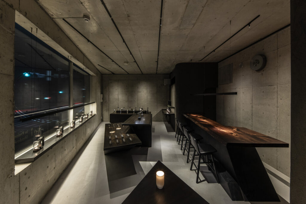 Candlelit interior with exposed concrete walls, a long wooden counter, and minimalist black display tables in Lei’s flagship store.