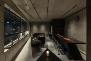 Candlelit interior with exposed concrete walls, a long wooden counter, and minimalist black display tables in Lei’s flagship store.