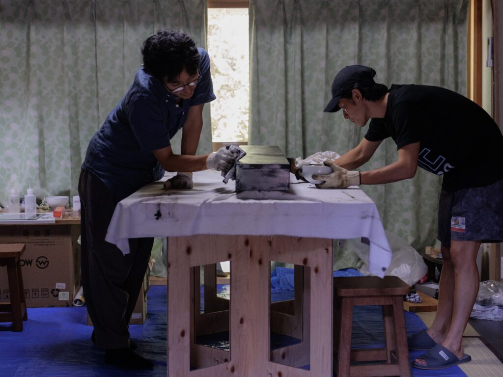 手袋をした2人が室内で木製パーツに拭き漆を施している作業風景