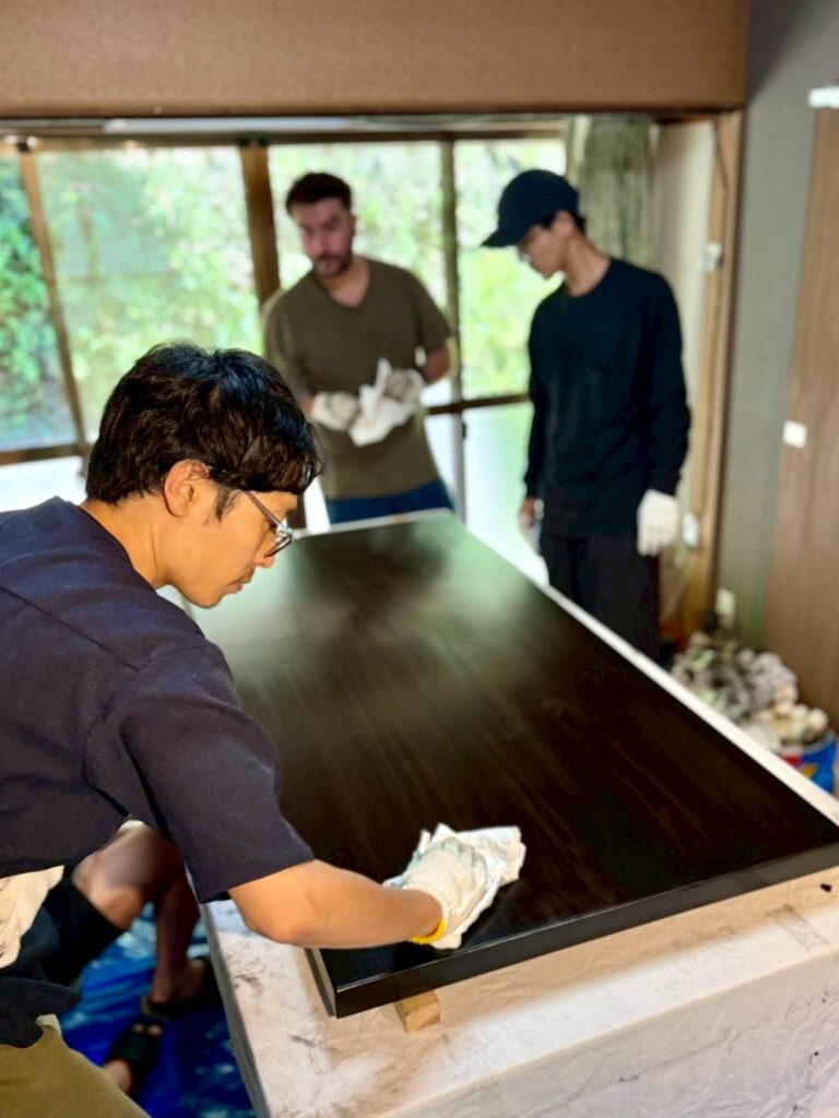 手袋をした職人(室瀬祐)が大きな黒い天板を布で拭き上げ、拭き漆で仕上げている作業風景