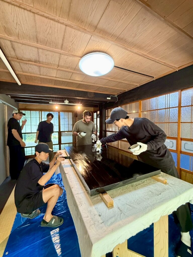 Team applying and inspecting urushi lacquer on a large display stand surface for Lei In Praise of Shadows Flagship Store, with one member photographing the process.