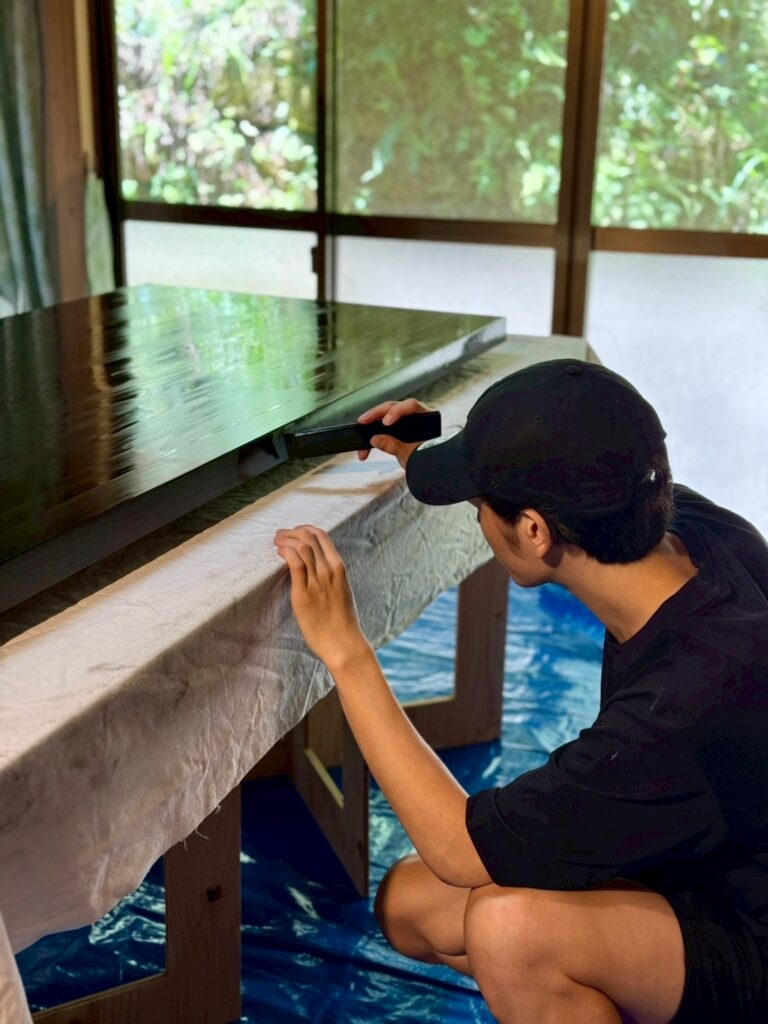 Leiブランドマネージャー佐藤が、が大きな黒い天板の縁を道具で整え、拭き漆の仕上がりを確認している様子