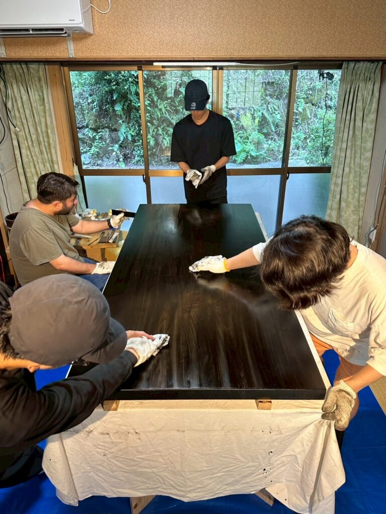 4人が手袋をして大きな黒い天板を囲み、布で拭き漆の仕上げ作業をしている室内の様子
