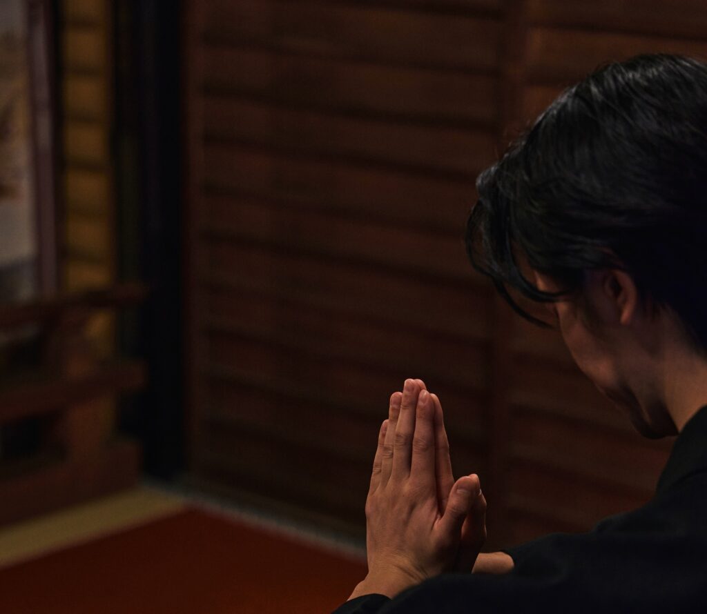 A person with hands pressed together in prayer inside a shrine, photographed from behind in warm, low light.