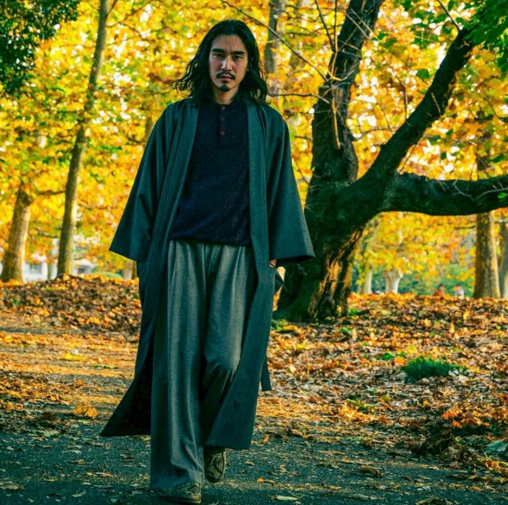 秋の木々の下、ローブのような羽織とワイドパンツを纏って並木道を歩く巧流-call-代表 元山巧大さん