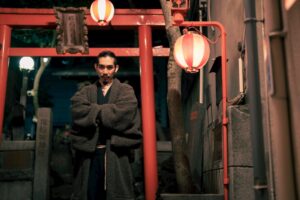 夜の鳥居の前で、羽織のような装いを纏った人物が腕を組んで立つ（提灯の灯り）