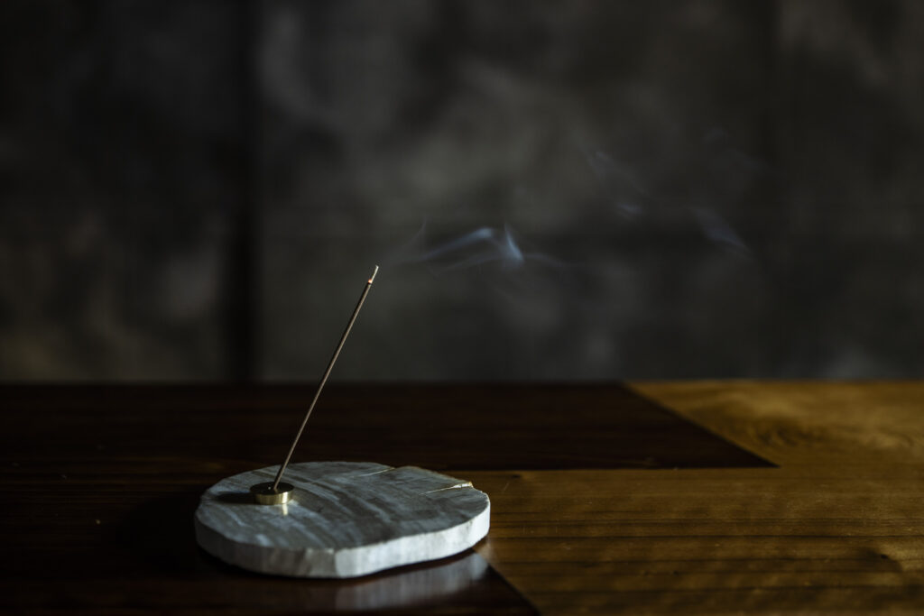 木のテーブルの上で燃える一本のお香。石の香立てから細い煙が立ちのぼり、暗い背景に静かに溶けていく。