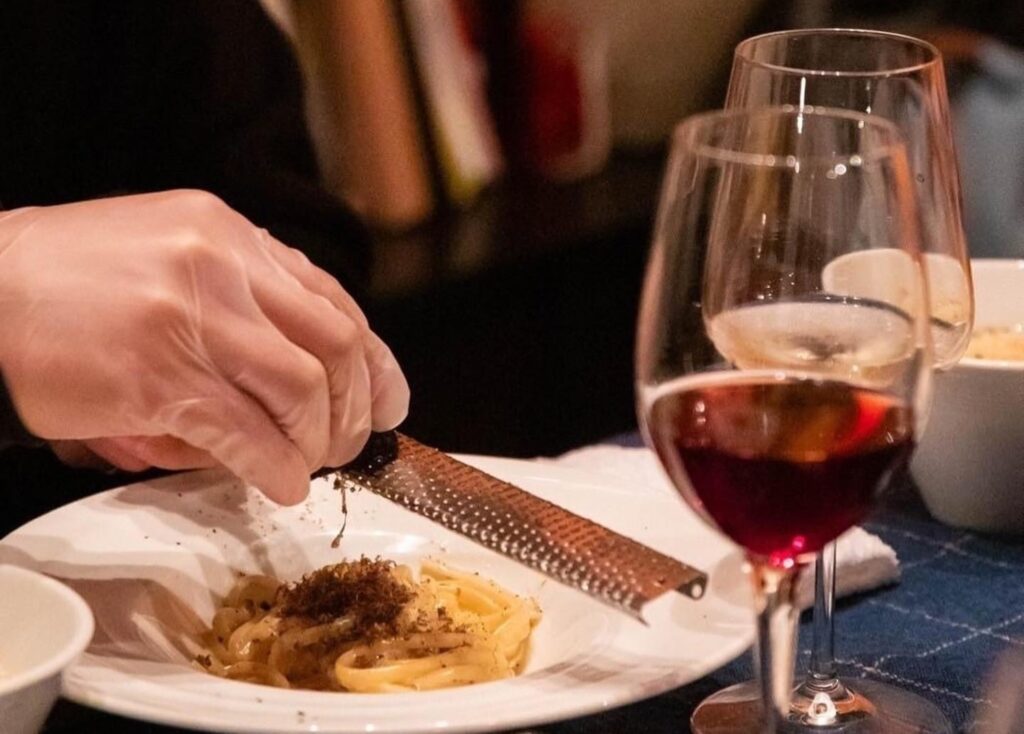 trattoria filoの料理。皿の上のパスタに、手元でトリュフ（またはチーズ）を削りかけて仕上げている場面。ワイングラスが並ぶ。