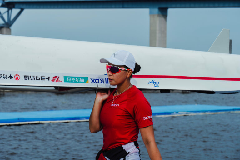 水辺で、ボート（艇）を肩に担いで歩く中条彩香さん。赤いシャツと白いキャップ、背後に橋。