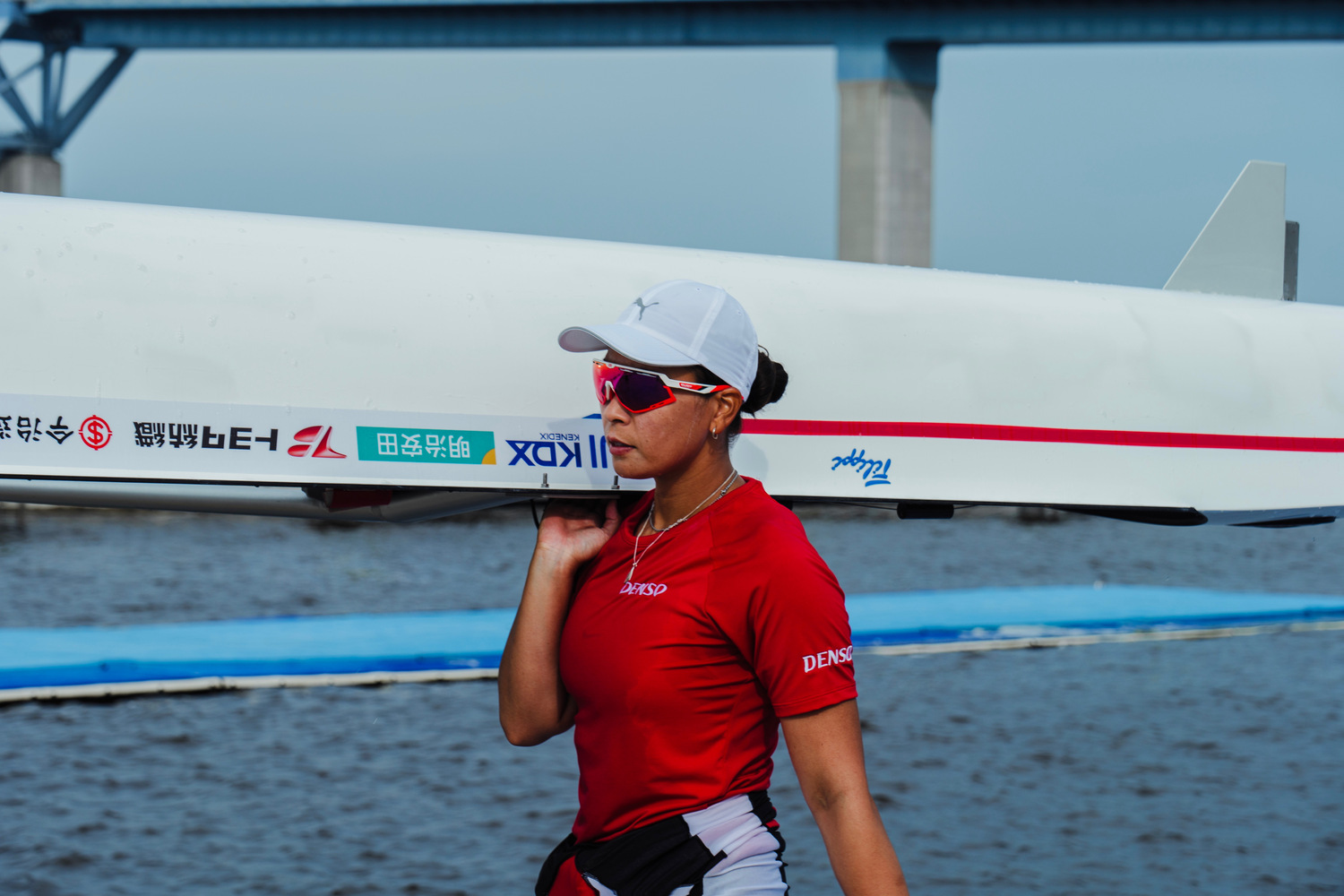 水辺で、ボート（艇）を肩に担いで歩く中条彩香さん。赤いシャツと白いキャップ、背後に橋。