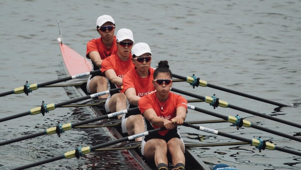 4人乗りのローイング艇で、赤いシャツのクルーが水面を進む。4本のオールが揃って伸びる。