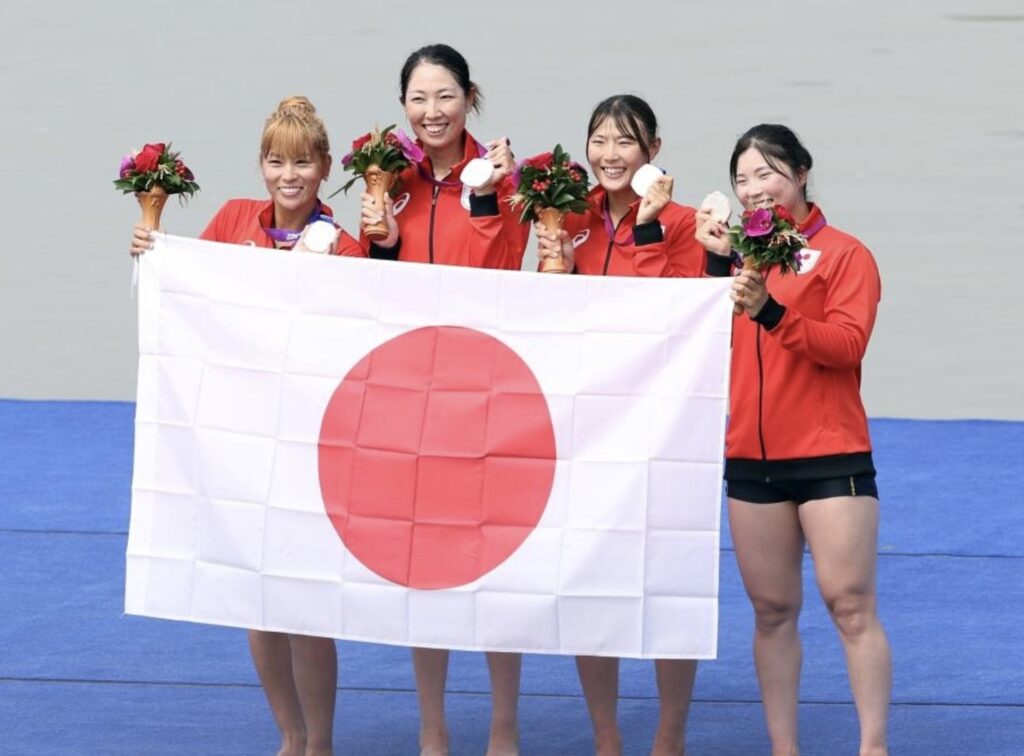 ローイング日本代表の4人の選手が、メダルと花束を手に日の丸を掲げて笑顔で並ぶ表彰シーン。