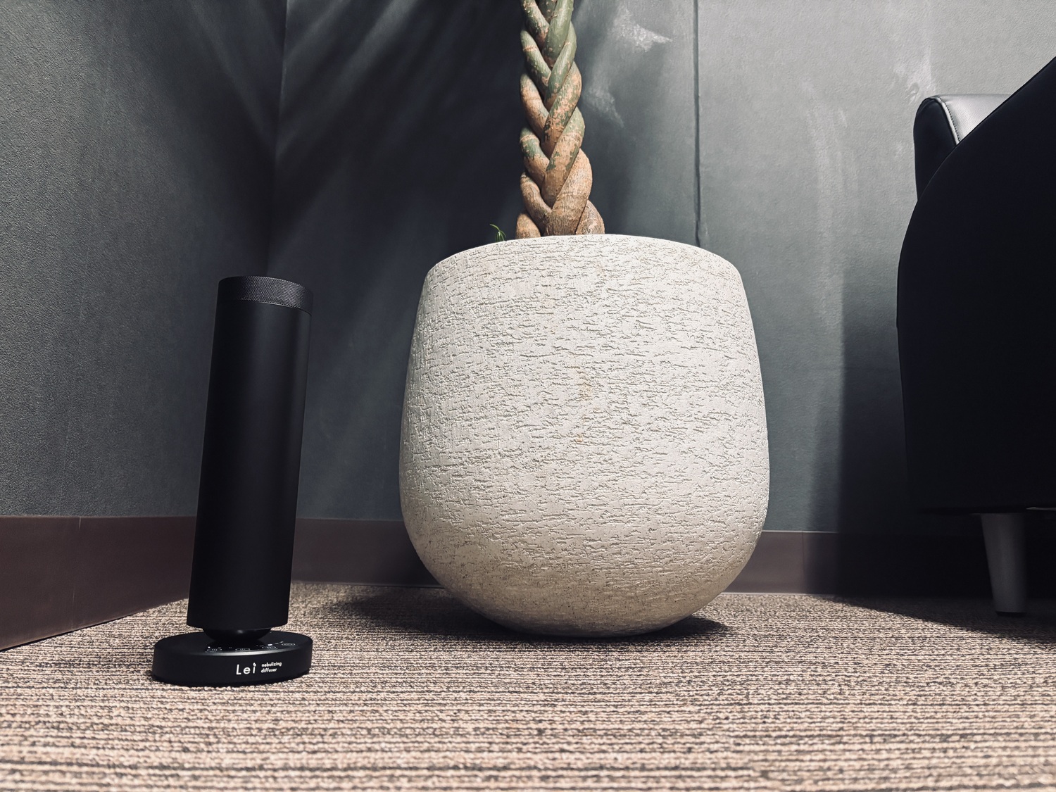 カーペットの床に置かれたLei業務用ネブライジングディフューザー。隣に大きな石調の鉢と観葉植物があるオフィスの一角。