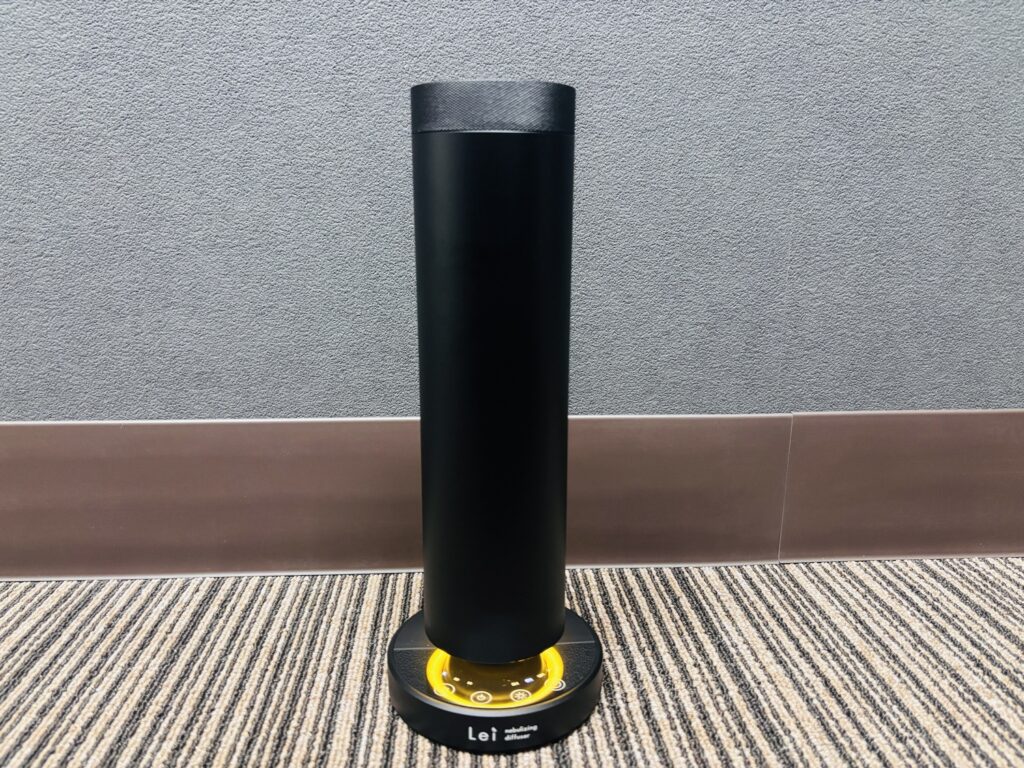 カーペットの床の上に設置されたLeiの業務用ネブライジングディフューザー。黒い筒型ボディと円形ベースが写っている。