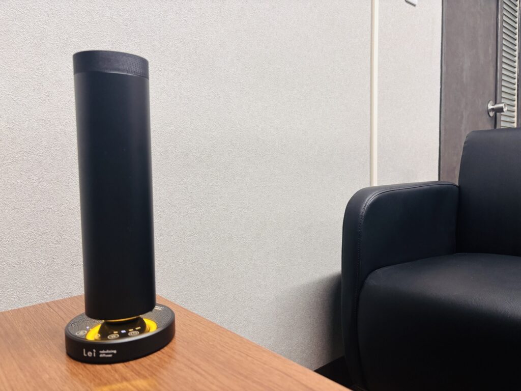 木製テーブルの上に置かれたLeiの業務用ネブライジングディフューザー。横に黒いソファがある打ち合わせスペース。
