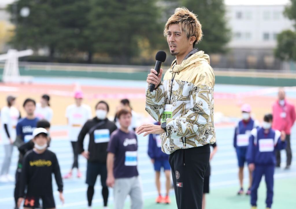 陸上競技場でマイクを手に話す藤光謙司さん。背後に参加者が並ぶ様子。