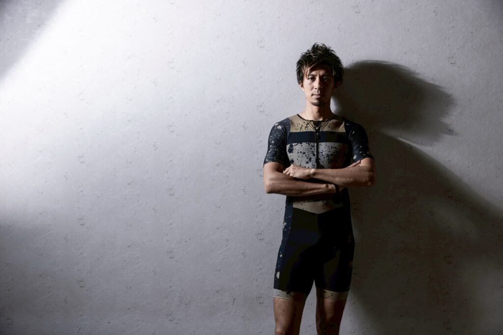 壁の前で腕を組んで立つ藤光謙司さん。強い光と影が落ちるポートレート。