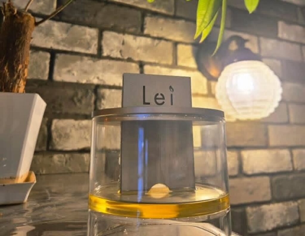 レンガ壁と柔らかな灯りの前に置かれた、Lei card diffuser（ガラス容器と香りのリフィル）