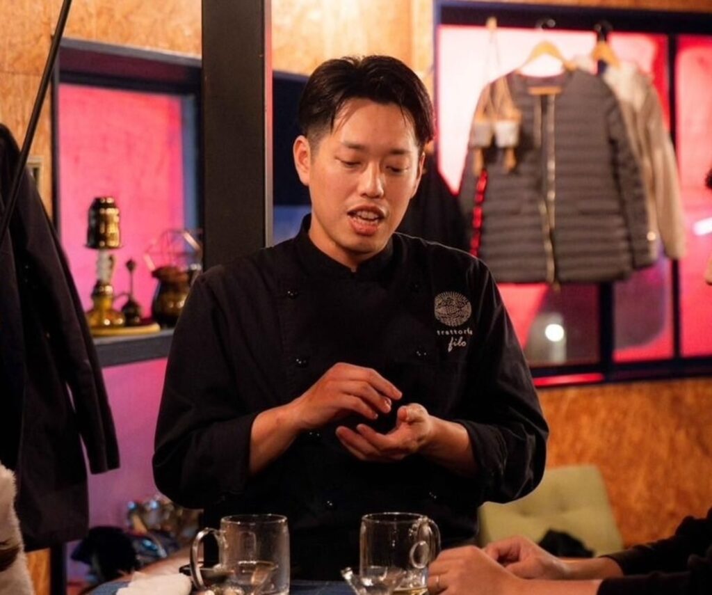 trattoria filoの店長兼シェフ・伊藤泰宏さん。黒いコックコート姿で、テーブルを囲みながら手元で説明している場面。