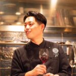 trattoria filoの店長兼シェフ・伊藤泰宏さん。厨房を背景に、ワイングラスを手に会話する様子。