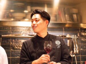 trattoria filoの店長兼シェフ・伊藤泰宏さん。厨房を背景に、ワイングラスを手に会話する様子。