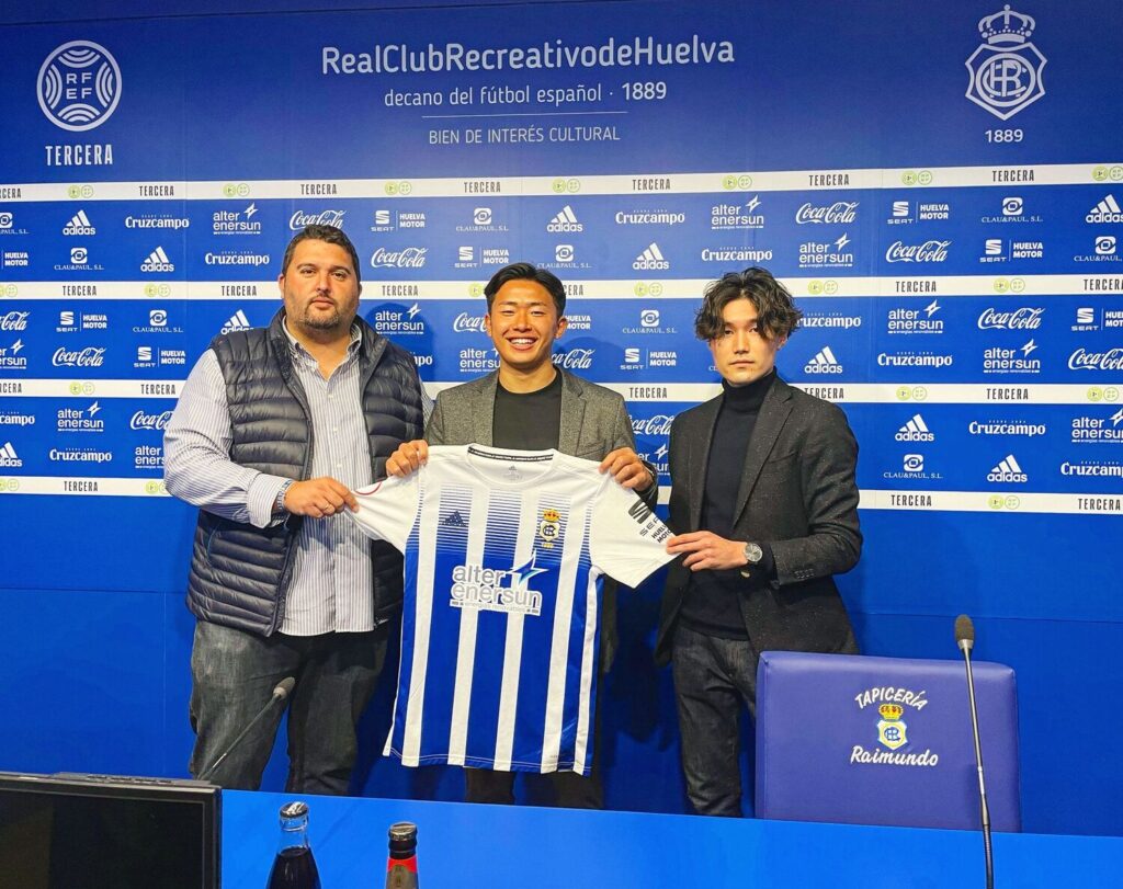 スペインのクラブ「Real Club Recreativo de Huelva」の記者会見会場で、3人がユニフォームを掲げて撮影している様子。