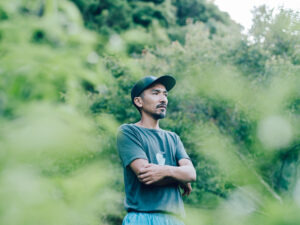 自然に囲まれた屋外で、腕を組みながら遠くを見つめる君島悠矢さんのポートレート