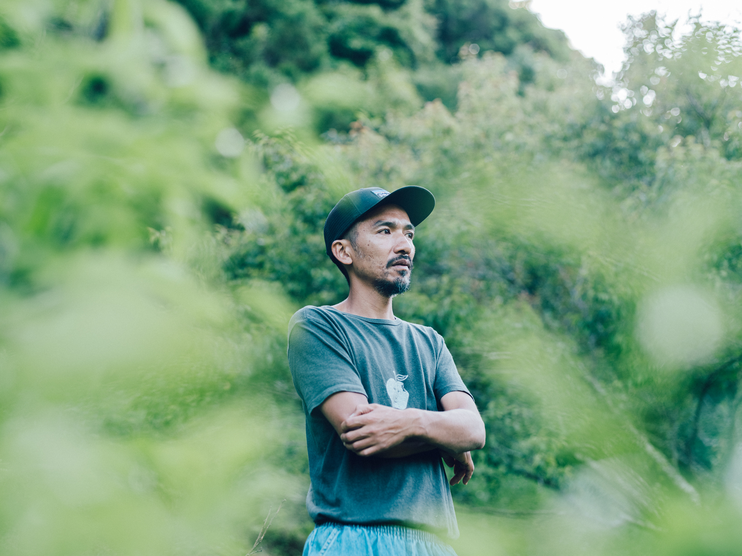 自然に囲まれた屋外で、腕を組みながら遠くを見つめる君島悠矢さんのポートレート