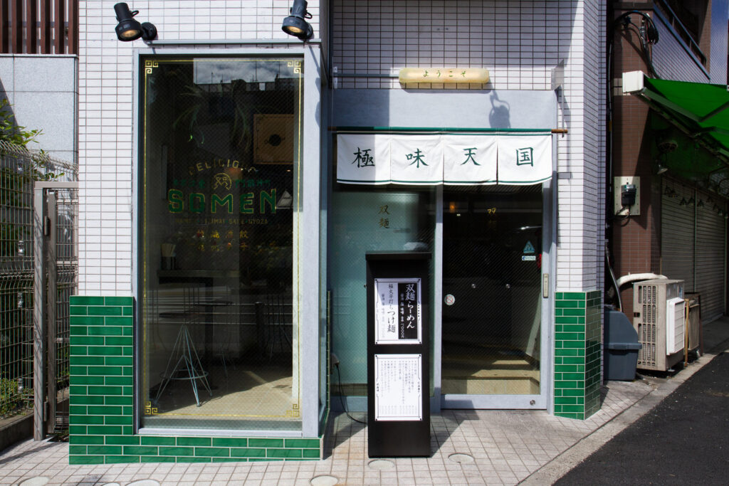 双麺の店舗外観。白い暖簾とガラス越しの店内、緑のタイルが印象的な昼の入口