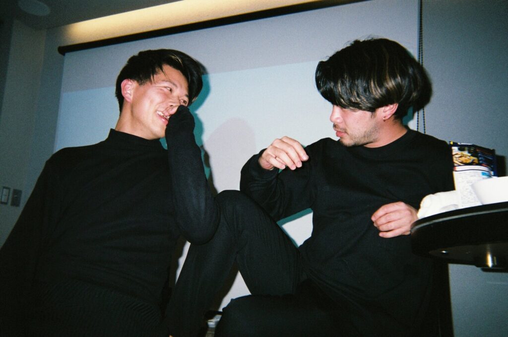 室内で笑いながら言葉を交わす塚本直純さんと男性のスナップ