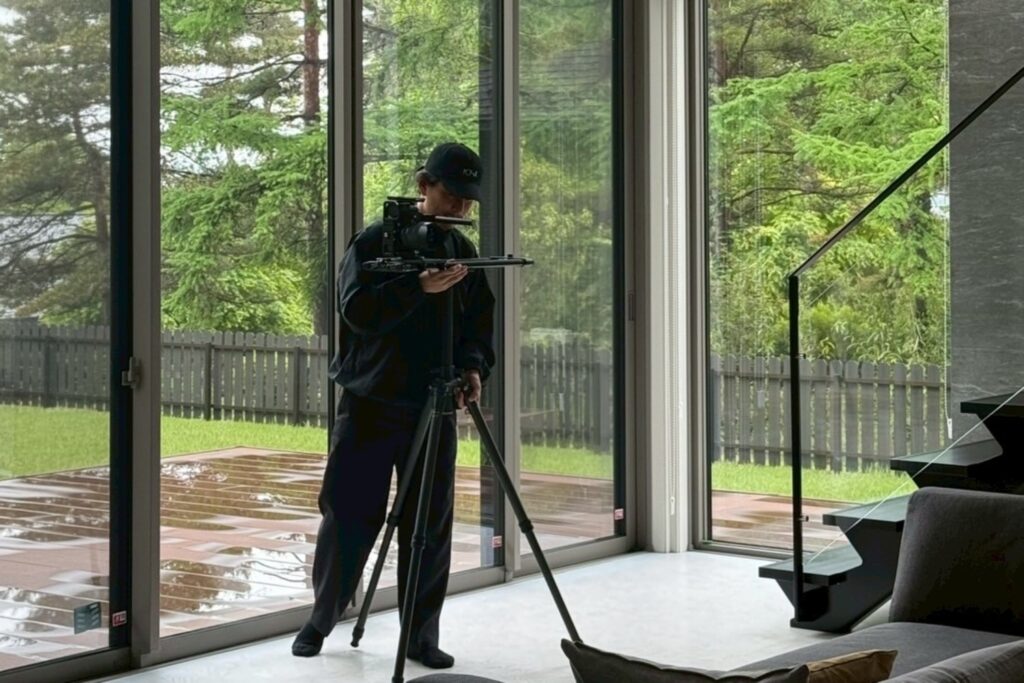 大きな窓辺の室内で三脚とカメラを調整する塚本直純さん