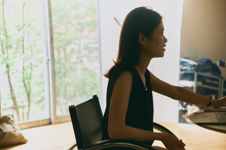 窓辺の自然光が差し込む室内で、椅子に座りながら話す藤井由香里さんの横顔