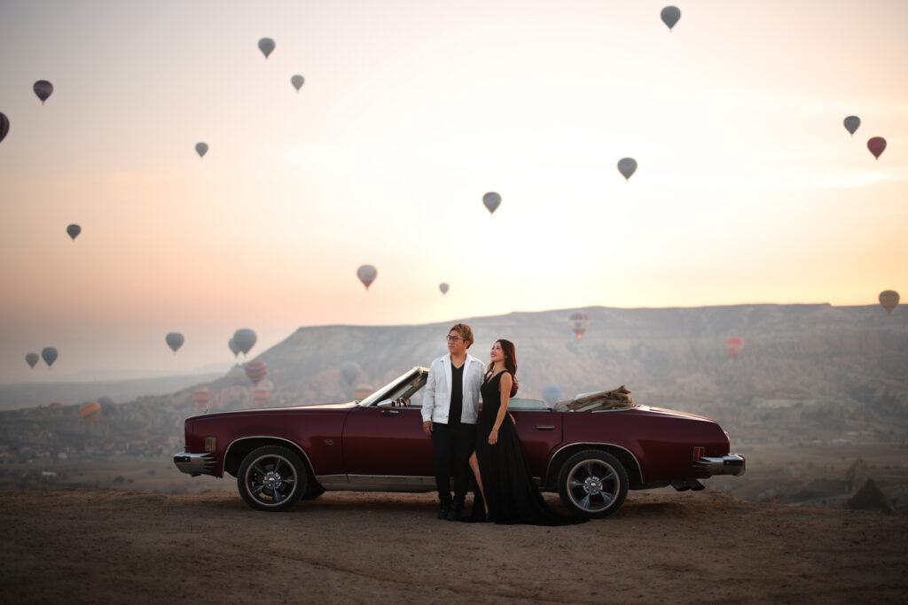 カッパドキアの夕景のなか、クラシックカーのそばに立つZEOさんとSUMMYさん。空には複数の気球が浮かんでいる。